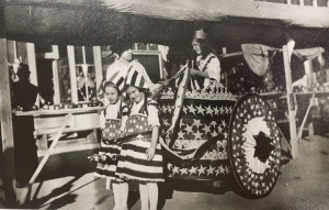 This photo taken during the 1915 Corn Carnival features Myrta Wagner Tway standing next to the chariot, holding the reins is Cataryne Atkinson, pulling the chariot are Helen Barto on left and Elizabeth Walters on the right. In the background is a block that burned down in the 1980s. (Modern day Love joys Plaza) 

Photo courtesy of the Plain City Historical Society. 